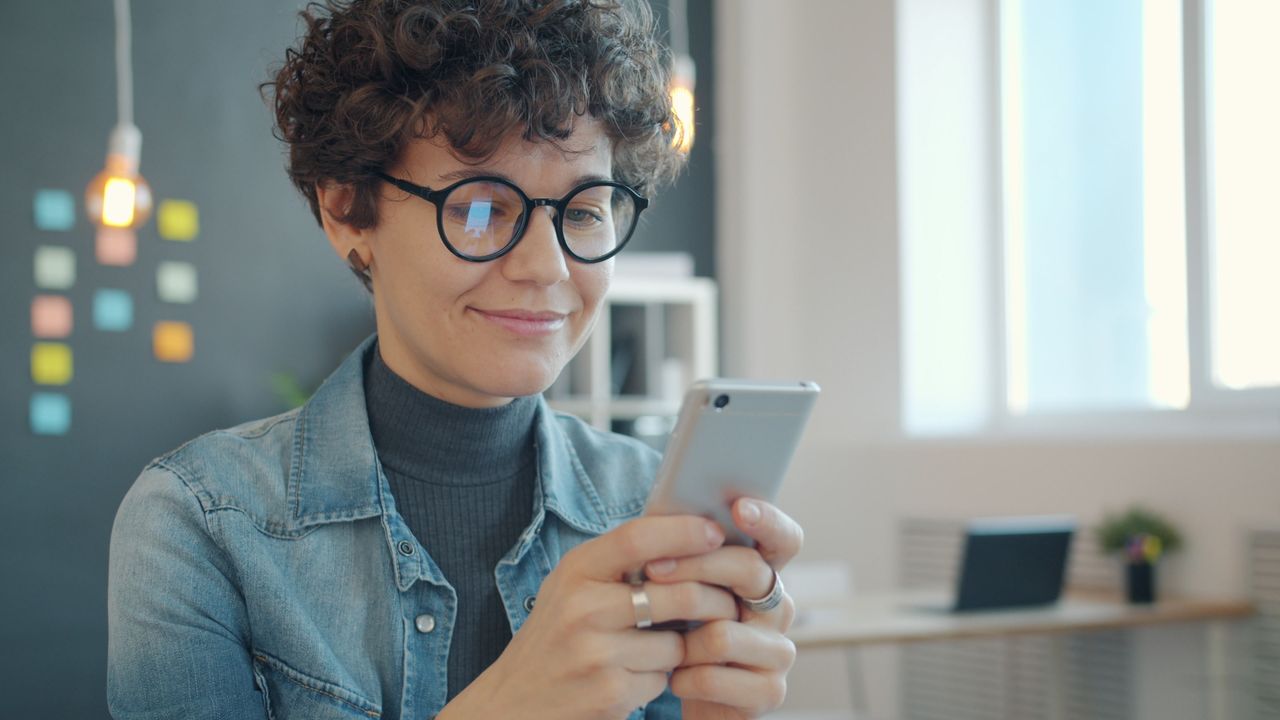 Services Woman in glasses using smartphone in modern office, smiling at screen.