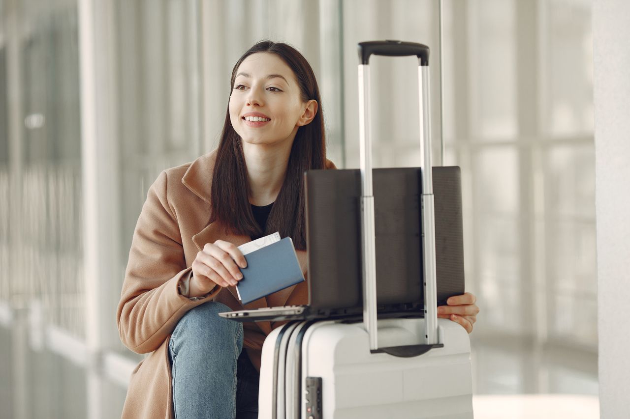 Services Smiling adult ethnic female traveler in trendy coat holding passport and using laptop on luggage in airport corridor for checking ticket for correctness