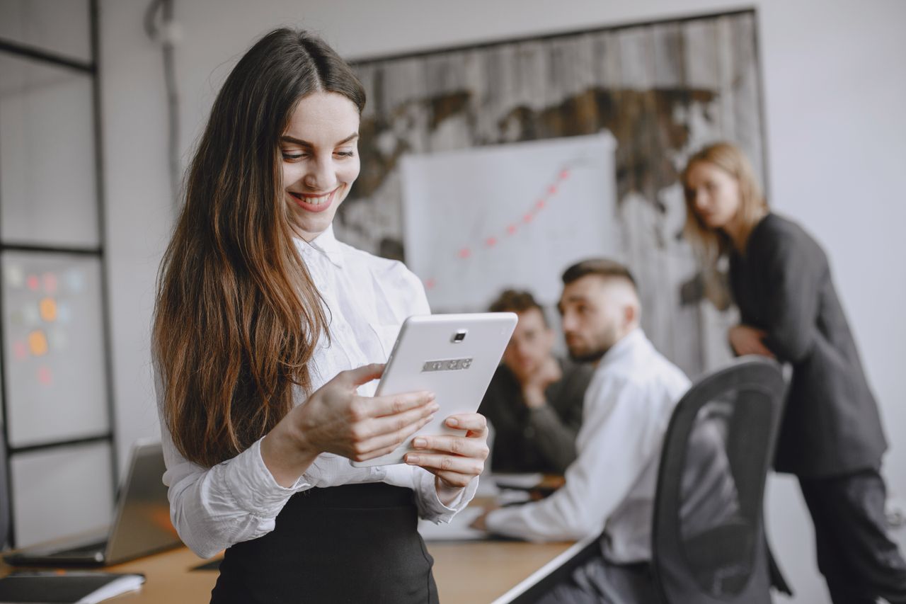 Services Young woman in a professional office setting browsing a tablet with colleagues in the background.